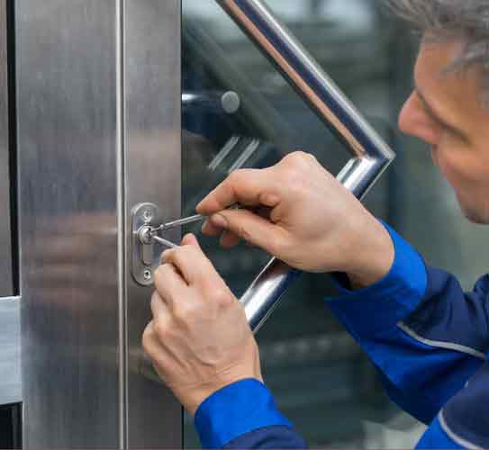 Locksmith holding tools in both hands to pick the lock of a glass door which has a metal frame. Represents Emergency Locksmith.
