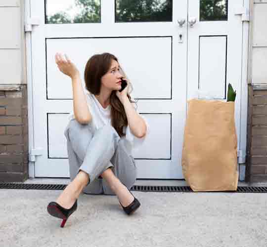 A woman sitting outside of a closed door with her shopping on the phone. Representing Locked out of your house and needing a locksmith.